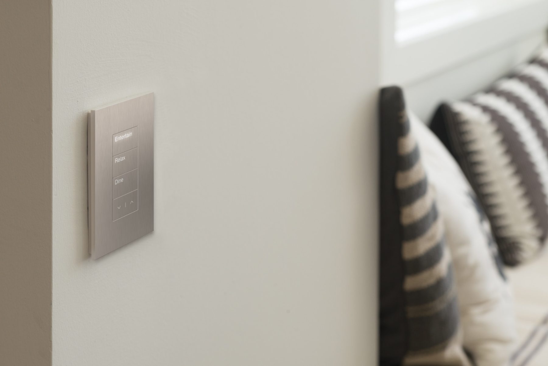 A brushed-metal Palladiom keypad engraved Entertain, Relax, Dine on a white wall beside a bed