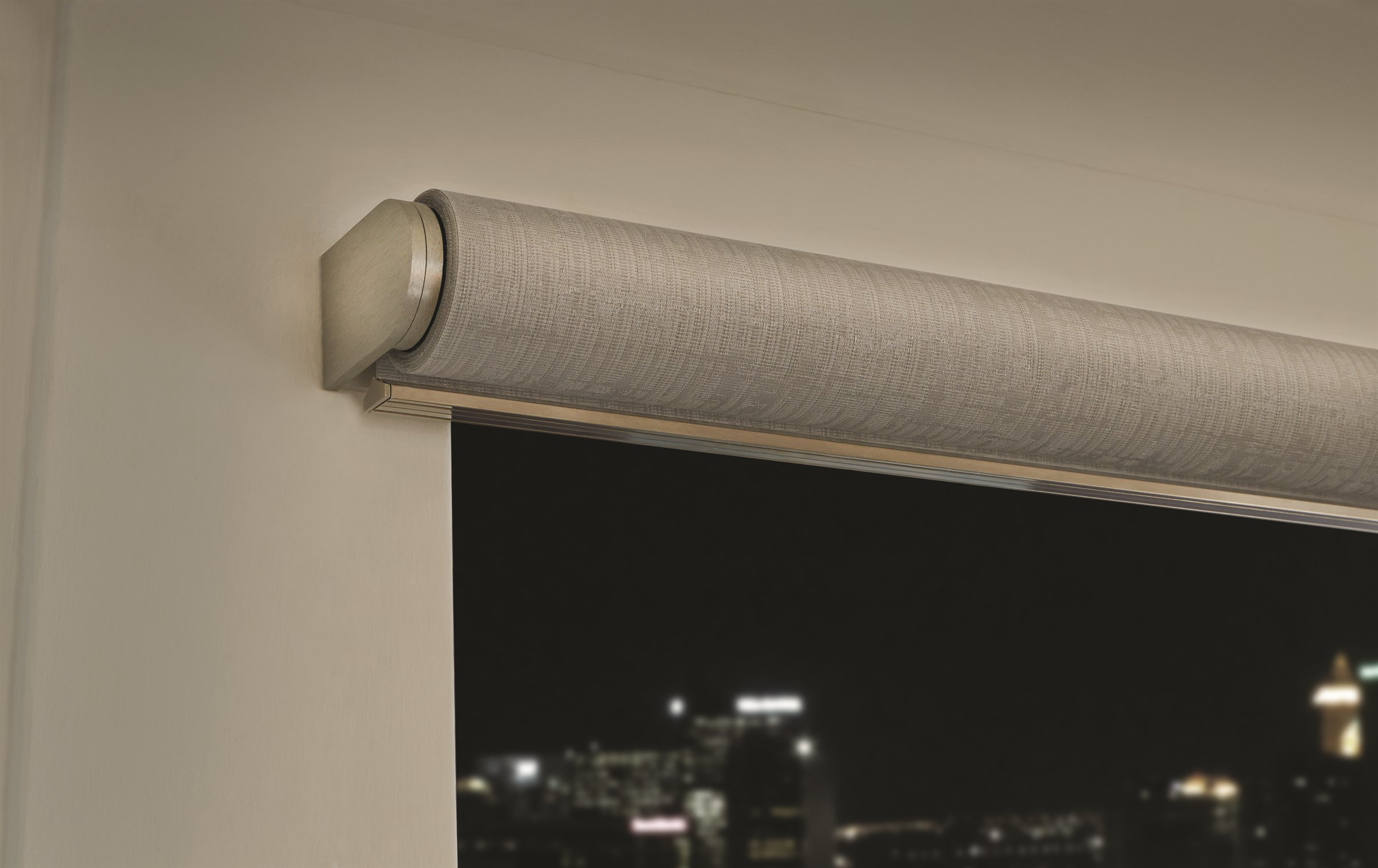 A Palladiom roller shade above a window, a city lit up at night beyond it